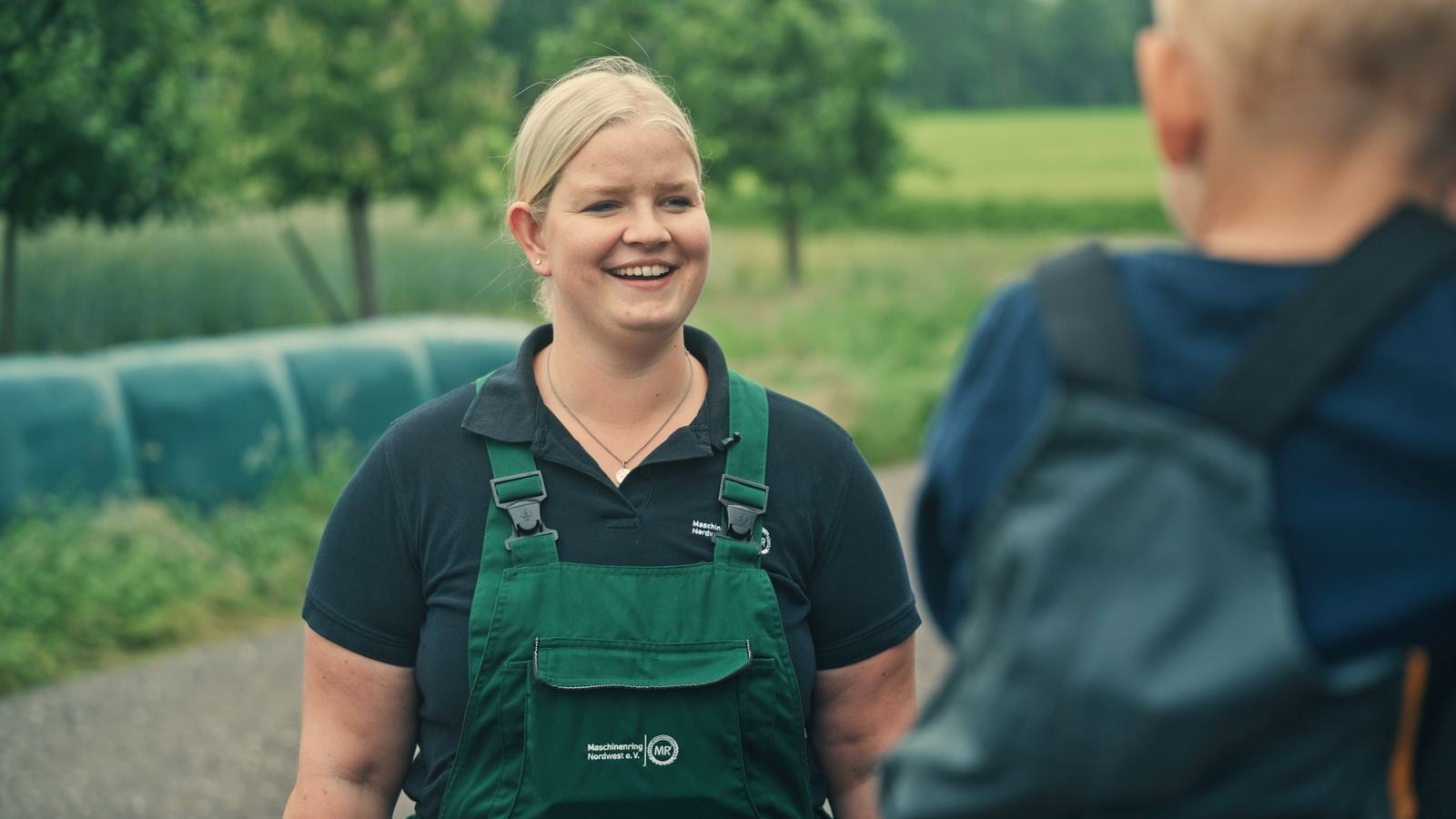 Lachende Betriebshelferin in grüner Latzhose im Gespräch auf dem Hof, im Hintergrund Siloballen