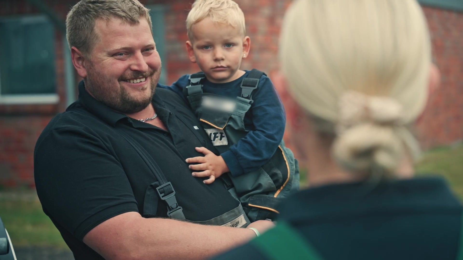 Landwirt mit seinem Sohn auf dem Arm lacht im Gespräch mit einer Betriebshelferin