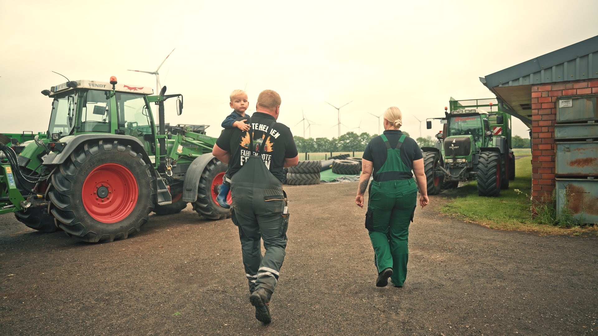 Landwirt mit seinem Sohn auf dem Arm und eine Betriebshelferin gehen über den Hof zu den Schleppern